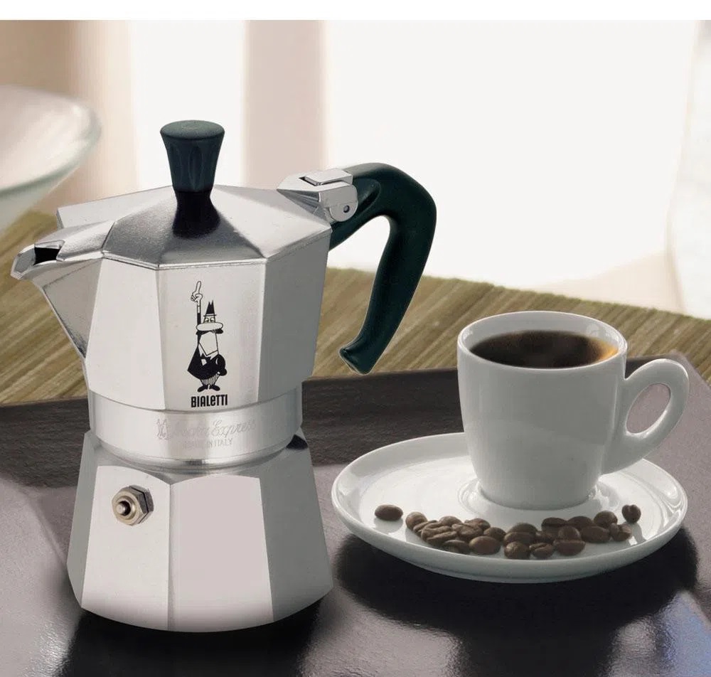 Cafeteira italiana na cor prata com xícara de café em cima de pires com alguns grãos de café em volta