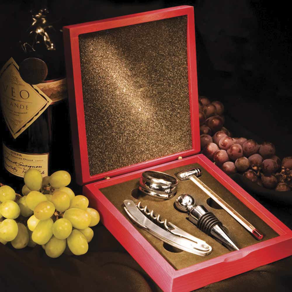 caixa aberta com kit para vinho contendo saca-rolha, colar, termômetro, tampa e uma caixa de madeira. Ao lado direito uva roxa, ao lado esquerdo uva verde e uma garrafa de vinho.