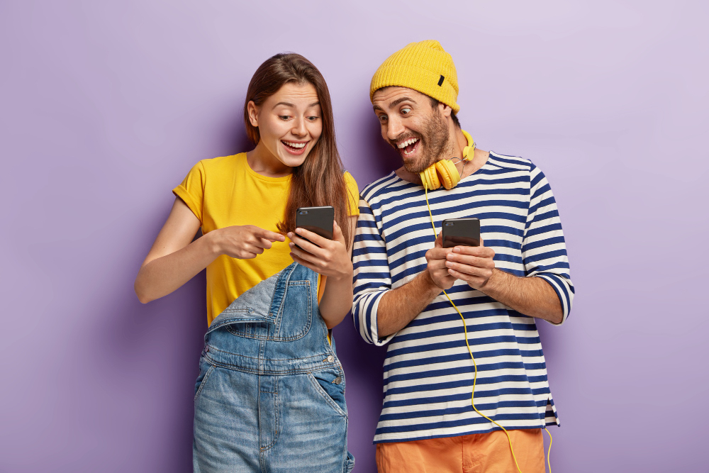 Do lado esquerdo, uma jovem mulher com celular na mão, sorrindo e olhando para o celular. Do lado aposto, jovem rapaz com celular na mão, sorrindo e olhando para o celular dela