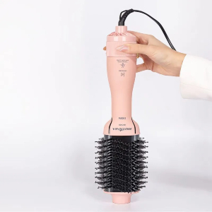 Mão feminina segurando escova secadora cor de rosa em cima de superfície branca.